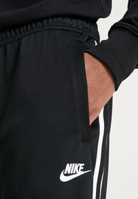 Person som bär svarta Nike jumpbyxor med vita sidostråk och elastisk midja, med handen delvis i högra fickan.