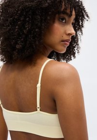 Νεαρή γυναίκα με σγουρά μαλλιά, που φορά ένα ανοιχτό κίτρινο, υφασμάτινο μπουστάκι, κοιτάζοντας πάνω από τον ώμο της σε μονόχρωμο φόντο.