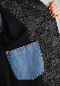 Kabát z vlněné směsi s texturovaným černobílým vzorem, s kontrastní modrou džínovou kapsou přišitou do vnitřního podšívky.