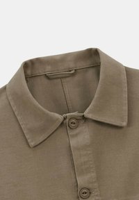 Chemise marron à col, avec deux boutons visibles sur la patte de boutonnage et des coutures renforcées autour du col et de la zone des boutons.