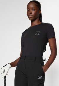 Zwarte katoenen t-shirt met een hart en gekruiste lijnen graphic in zilver, gecombineerd met zwarte broek met een EA7-logo. Textured handschoen en golfclub zichtbaar.