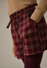 Pantaloni corti a scacchi bordeaux con accenti rosa, dotati di una vita con lacci e tasche laterali. Abbinati a collant a costine color bordeaux.