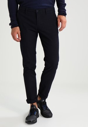 Man draagt strakke zwarte broek, zwarte textuur sneakers met blauwe accenten, en een donker shirt met lange mouwen, staand op een witte achtergrond.
