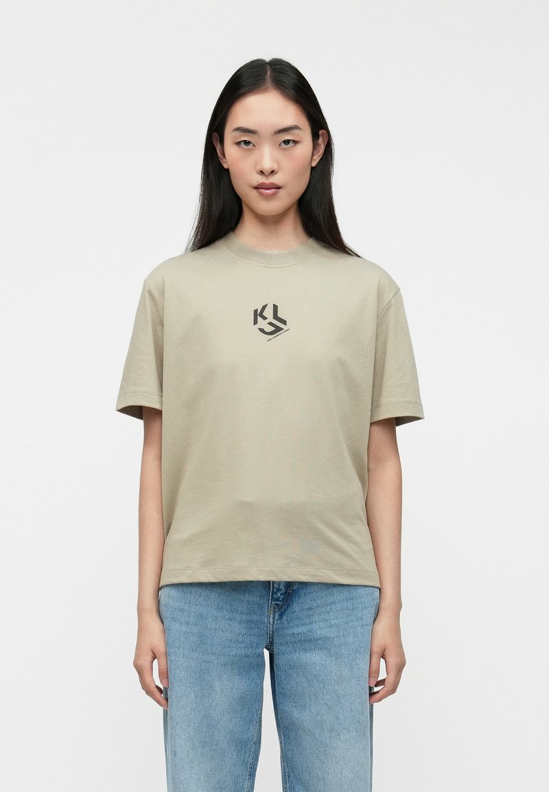 Kaki T-shirt med korte ærmer, rund hals og et sort logo på brystet; båret med blå denimjeans. Glat bomuldsstof.