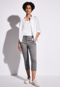 Femme se tenant contre un mur blanc, portant une chemise blanche, un débardeur blanc, un pantalon gris court et des baskets blanches, avec une main dans la poche.