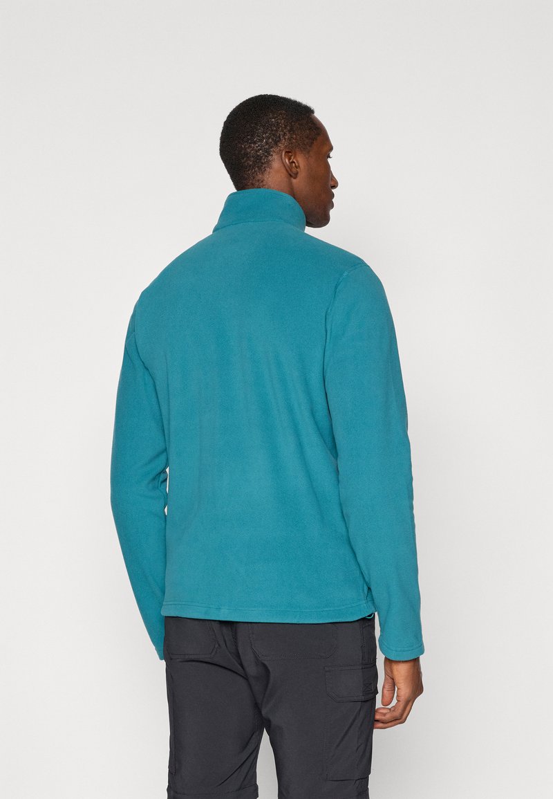 Turkusowa polarowa kurtka z wysokim kołnierzem, długimi rękawami i gładką teksturą, zestawiona z ciemnymi spodniami cargo. Widoczne detale szycia na końcach.