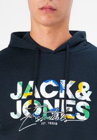 Marineblaue Kapuzenjacke mit Kordelzug, ausgestattet mit einem großen, mehrfarbigen „JACK & JONES Essentials“ Grafikdruck in Weiß, Blau, Grün und Gelb.