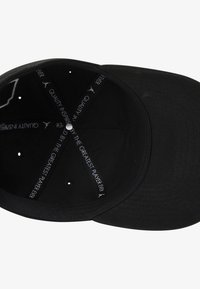 Svart baseballcaps med flat skygge, tekstur av stoff og innvendig detaljer med hvit tekst som sier "Kvalitet inspirert av den største spilleren noensinne."