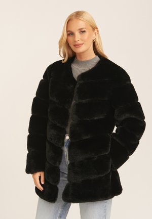 Black faux fur coat with a puffer design, features horizontal seams, a rounded collar, and long sleeves. Worn over a gray top and denim.