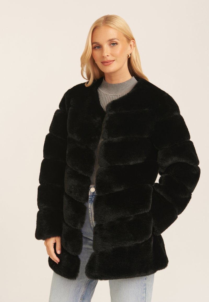 Black faux fur coat with a puffer design, features horizontal seams, a rounded collar, and long sleeves. Worn over a gray top and denim.