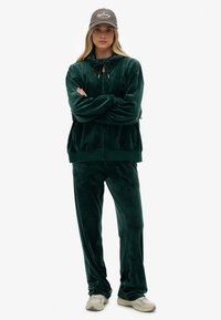 Grön velour joggingdress med huva, dragkedja och matchande byxor. Har snörning, broderad logga och en mjuk yta.