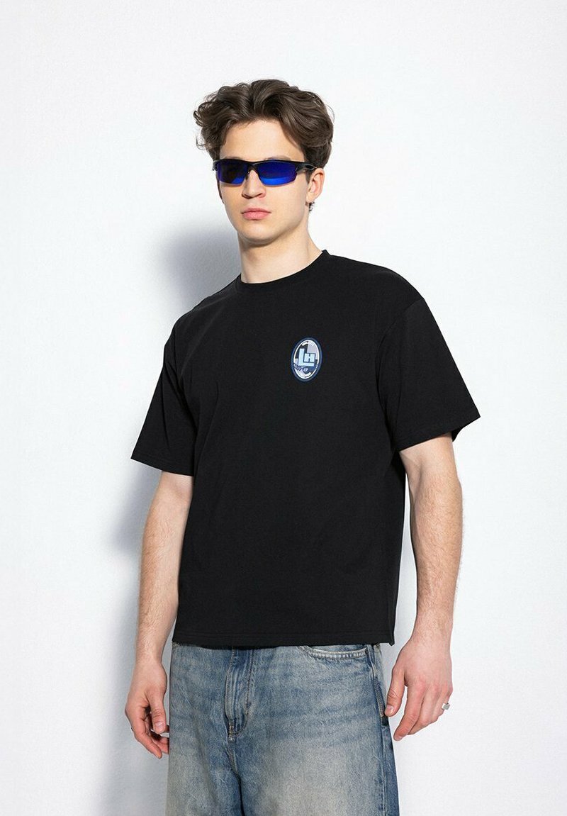 Czarna bawełniana koszulka z okrągłym dekoltem i krótkimi rękawami, z małym niebiesko-białym owalnym logo na klatce piersiowej. Noszona z niebieskimi okularami przeciwsłonecznymi.