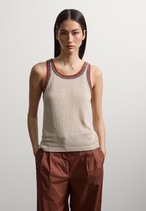 Jonge vrouw met stijl donker haar die een beige gebreide tanktop met multicolor bies en bruine hoog getailleerde broek draagt, handen in haar zakken.