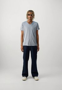 Svetlo modra bombažna majica z V-izrezom, kombinirana s temno modrimi kavbojkami z širokimi hlačnicami in belimi supergami. Doplnjena s sončnimi očali.