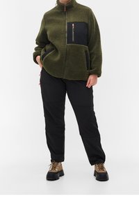 Giacca in fleece verde oliva con tasca sul petto con zip nera e tasche laterali, abbinata a pantaloni neri e stivali marroni con lacci.