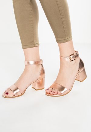Voeten met rosegouden sandalen met blokhak en enkelbandjes, gecombineerd met cropped beige broek, op een witte achtergrond.