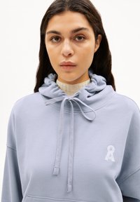 Lichtblauwe hoodie van zacht materiaal, met een trekkoord in de capuchon en een buidelzak aan de voorkant, en een klein wit logo op de borst.