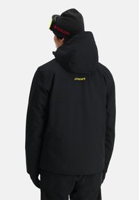 Schwarze wasserdichte Jacke mit Kapuze, mit einem gelben "SPYDER"-Logo auf dem Rücken. Glatte Textur, minimalistisches Design, geeignet für Outdoor-Aktivitäten.