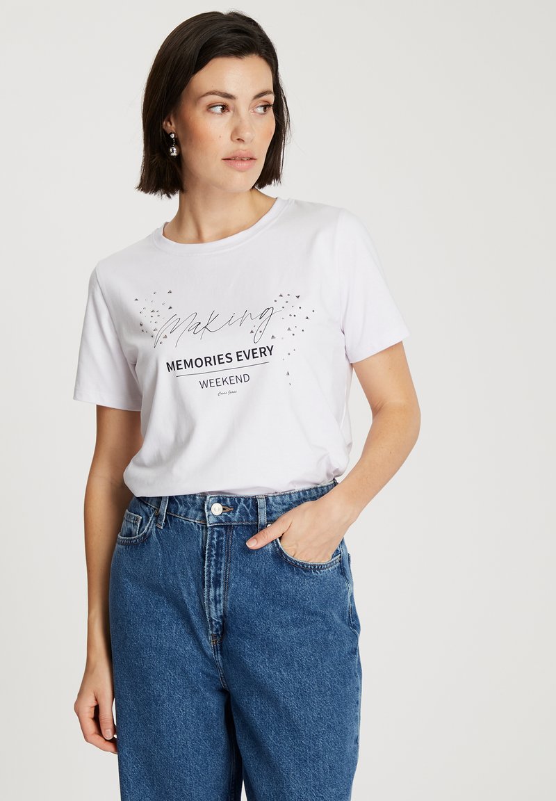Biała bawełniana koszulka z czarnym napisem „Tworzymy Wspomnienia Każdego Weekendu” z błyszczącymi akcentami, w połączeniu z jasnoniebieskimi dżinsami o podwyższonym stanie.