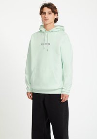 Mintgroene hoodie met een kangoeroezak, trekkoorden en geborduurd "VOLCOM"-logo in paars. Combinatie met zwarte broek. Zachte textuur.