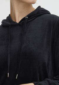 Crna hoodie od mekog, plišanog materijala s ležernim krojem, koja ima kapuljaču s uvezenom žicom i metalnim vrhovima. Glatka tekstura i jednostavan dizajn.