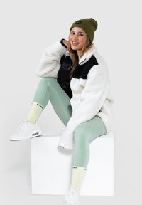 Giacca di pile bianca con accenti neri, leggings verde chiaro, sneakers bianche e berretto verde oliva. La texture è morbida e stratificata.