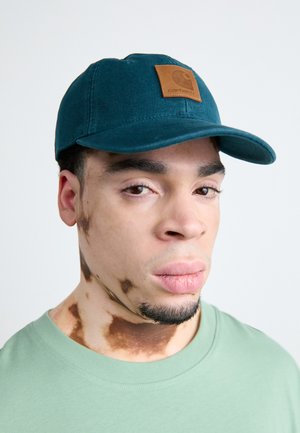 Türkise Baumwollmütze mit einem braunen Lederpatch, auf dem ein Logo zu sehen ist. Modell trägt ein hellgrünes T-Shirt, das eine glatte Textur zeigt und vor einem neutralen Hintergrund präsentiert wird.