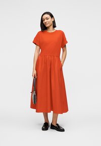 Κόκκινο βαμβακερό φόρεμα με κοντά μανίκια, στρογγυλή λαιμόκοψη και φούστα που φαρδαίνει. Διαθέτει σφηκοειδή μέση. Συνδυάζεται με μαύρα μοκασίνια και τσάντα.