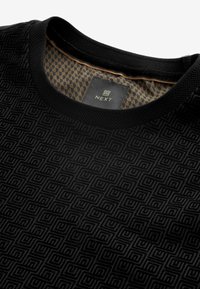 Camisa negra de manga corta con un patrón geométrico texturizado. Cuello redondo con un suave collar negro y un forro interno de patrón contrastante.