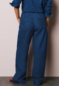 Tumši zili džinsa bikses ar elastīgu jostasvietu un divām sirsniņu formas aizmugures kabatām. Tām ir taisnu staru dizains un minimālas šuves.