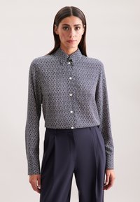 Camicia blu navy con un motivo geometrico bianco, maniche lunghe e colletto classico, abbinata a pantaloni scuri su misura.