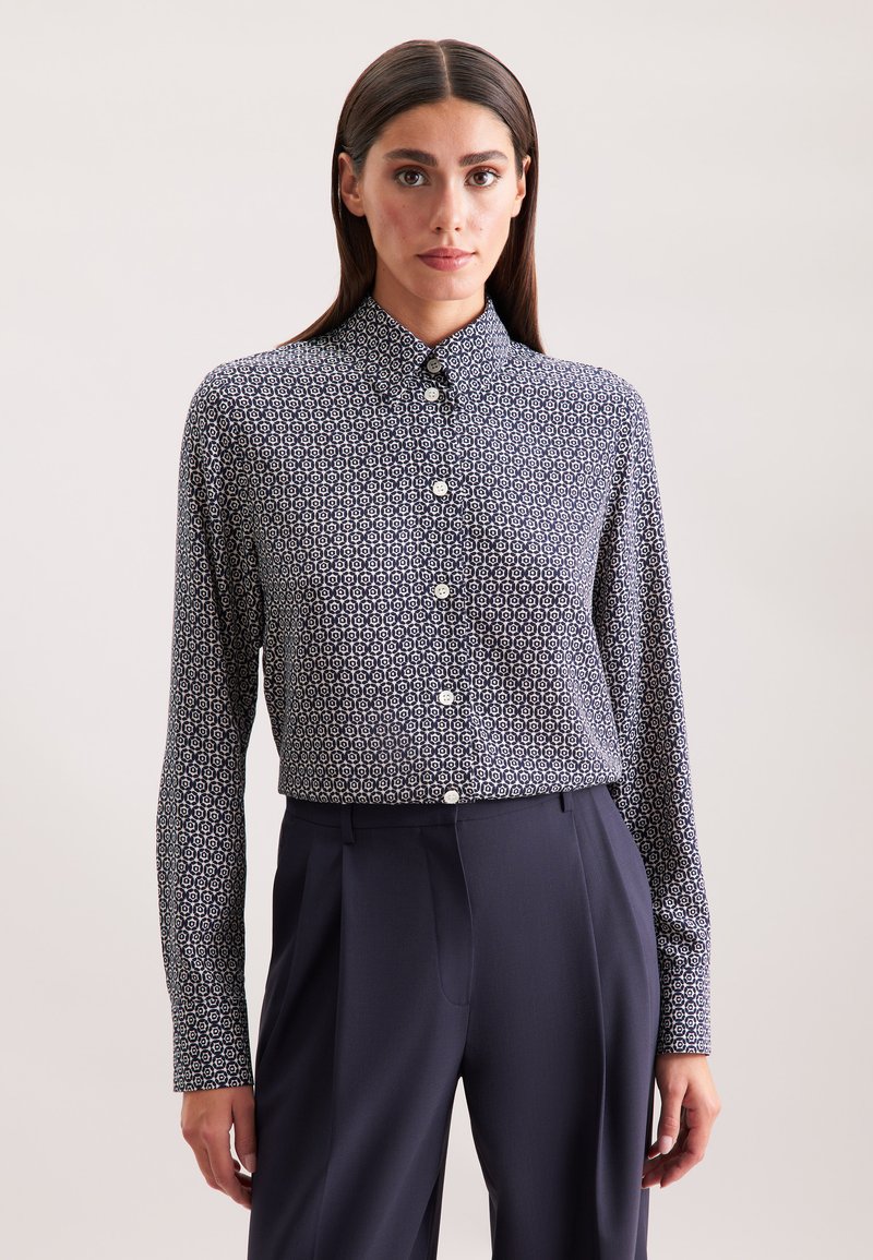 Camicia blu navy con un motivo geometrico bianco, maniche lunghe e colletto classico, abbinata a pantaloni scuri su misura.