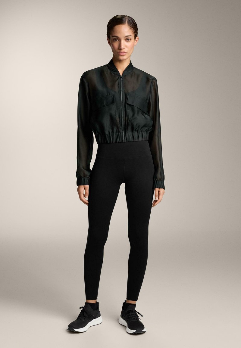 Musta läpikuultava bomber-takki, jossa on kaksi etutaskua, kiristetty vyötärö ja pitkät hihat, yhdistettynä korkeavyötäröisiin mustiin leggingsseihin ja mustiin lenkkareihin.