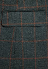 Tkanina wełniana w ciemnozielonym kolorze z delikatnym wzorem w kratę w odcieniu pomarańczowym. Zawiera fałszywą kieszeń i ma teksturowaną powierzchnię.