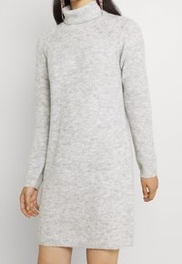 Abito maglione grigio chiaro a costine con collo alto, maniche lunghe e lunghezza leggermente sopra il ginocchio. Morbida texture, design minimal.