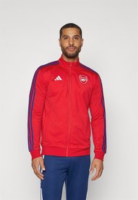 Rød atletisk jakke med blå striber på ærmerne, fuld lynlås foran, høj krave og Arsenal-logo på venstre bryst. Glat stof, tætsiddende design.