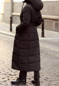 Schwarzer Steppmantel mit Kapuze, mit gesteppter Optik, langer Länge, elastischer Taille und Seitentaschen, kombiniert mit schwarzen Schnürstiefeln.