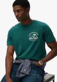 Zielony bawełniany t-shirt z grafiką góry, połączony z niebieską koszulą w kratę zawiązaną w talii oraz dżinsami. Złoty zegarek na nadgarstku.