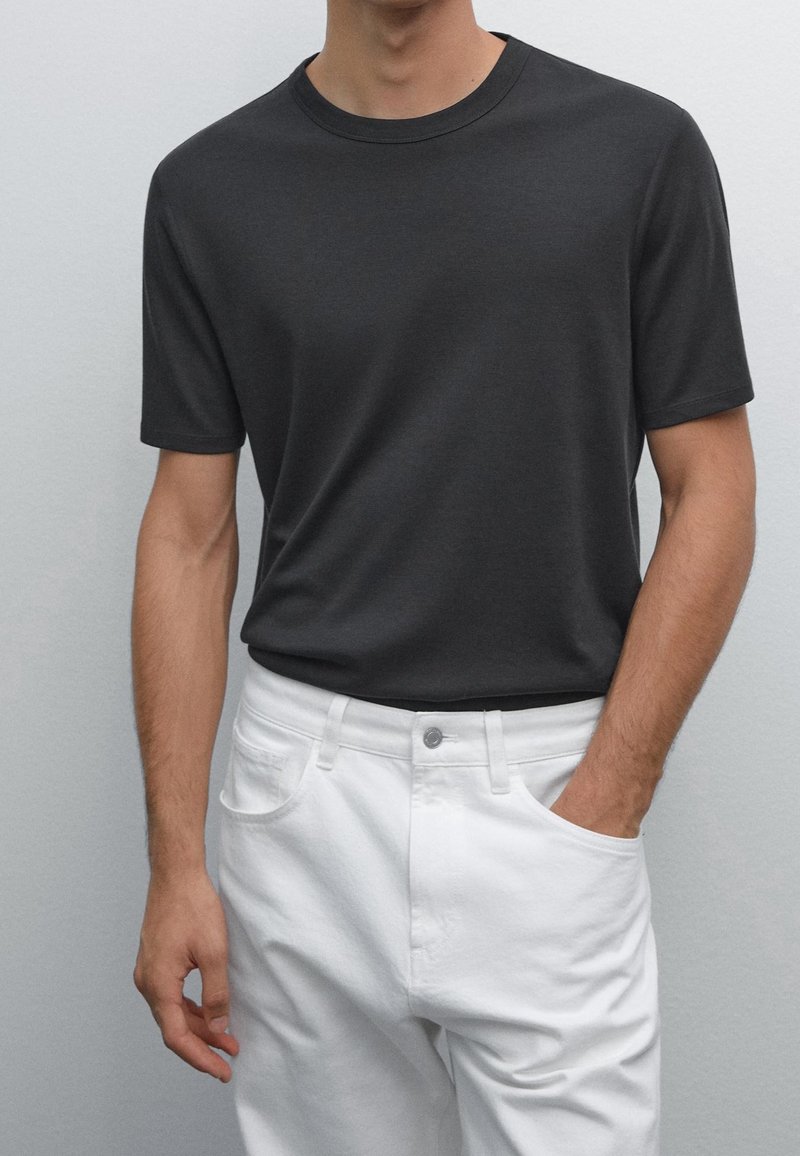 Homme portant un t-shirt gris foncé uni à manches courtes rentré dans un pantalon blanc avec une main dans la poche sur un fond uni.