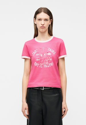 Γυναίκα που φοράει ροζ t-shirt με λευκή διακόσμηση, με ένα κινούμενο ποντίκι σε σκέιτμπορντ και λευκό κείμενο, συνδυασμένο με μαύρο παντελόνι.