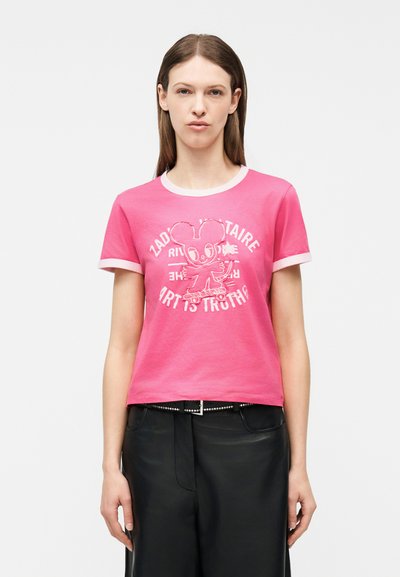 Γυναίκα που φοράει ροζ t-shirt με λευκή διακόσμηση, με ένα κινούμενο ποντίκι σε σκέιτμπορντ και λευκό κείμενο, συνδυασμένο με μαύρο παντελόνι.