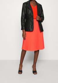 Zwarte leren blazer, oranje V-hals top, geplisseerde koraalkleurige rok en zwarte open teen hakken. Gestructureerd ontwerp met een mooie textuur.