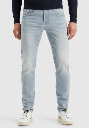 Light blue denim jeans with a slim fit, featuring subtle fading, five pockets, and metal hardware. Paired with white and tan sneakers.