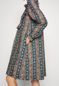 Květinové šaty ve tmavé modrozelené barvě s dlouhými rukávy, volánkovým límcem a kapsami. Mají živý květinový vzor v růžových a červených tónech.