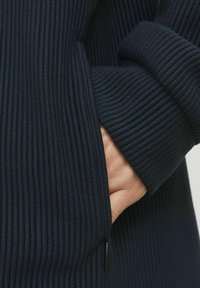 Marineblauer gerippter Stoff mit hohem Kragen und langen, umgeschlagenen Ärmeln. Eine Hand ist in die Tasche gesteckt, was die Textur und das weiche Material zur Geltung bringt.