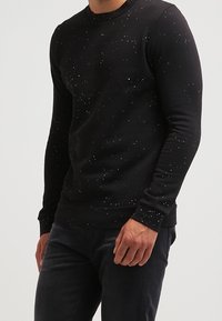 Uomo che indossa un maglione nero a maniche lunghe con un motivo screziato bianco, abbinato a pantaloni scuri, su uno sfondo semplice.
