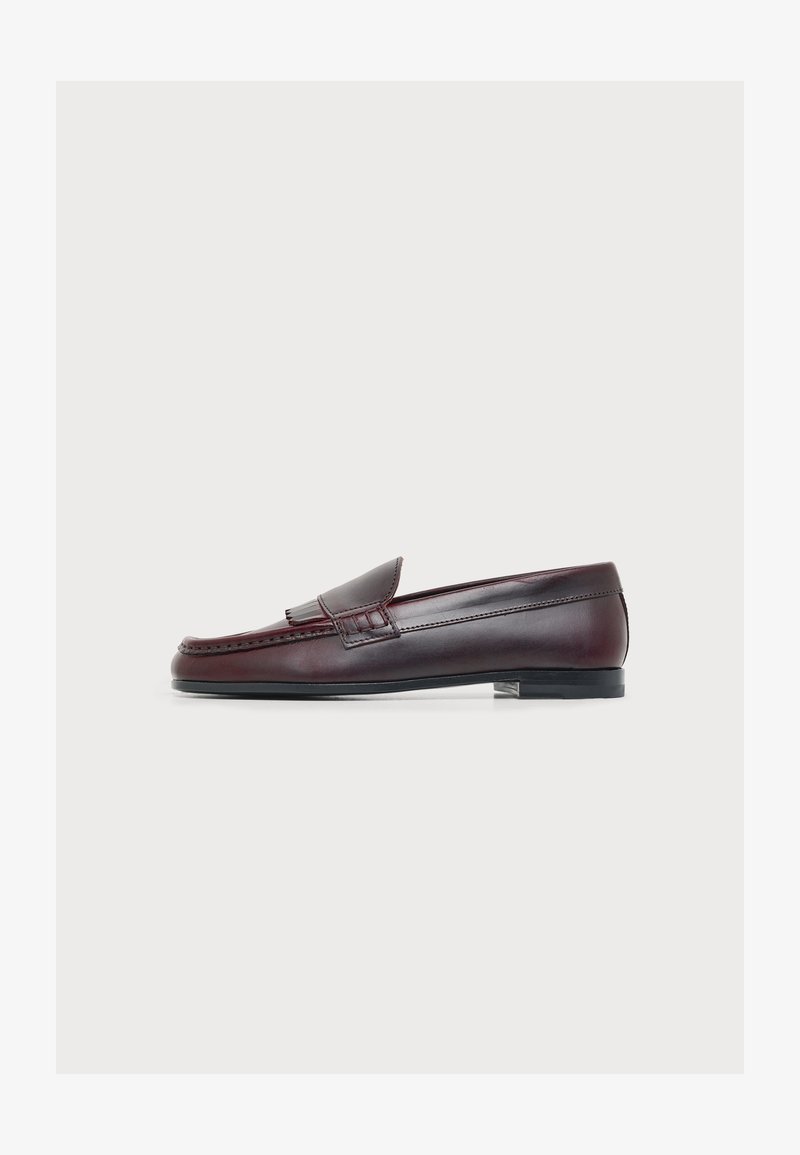 Mocassin en cuir marron foncé avec un petit talon, un bout arrondi et une frange décorative sur le dessus, sur fond blanc.