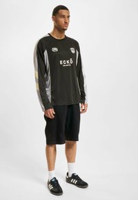 Svart langermet sportskjorte med grå og gull aksenter, med logoen "ECKO UNLIMITED" på forsiden, kombinert med svarte shorts og joggesko.