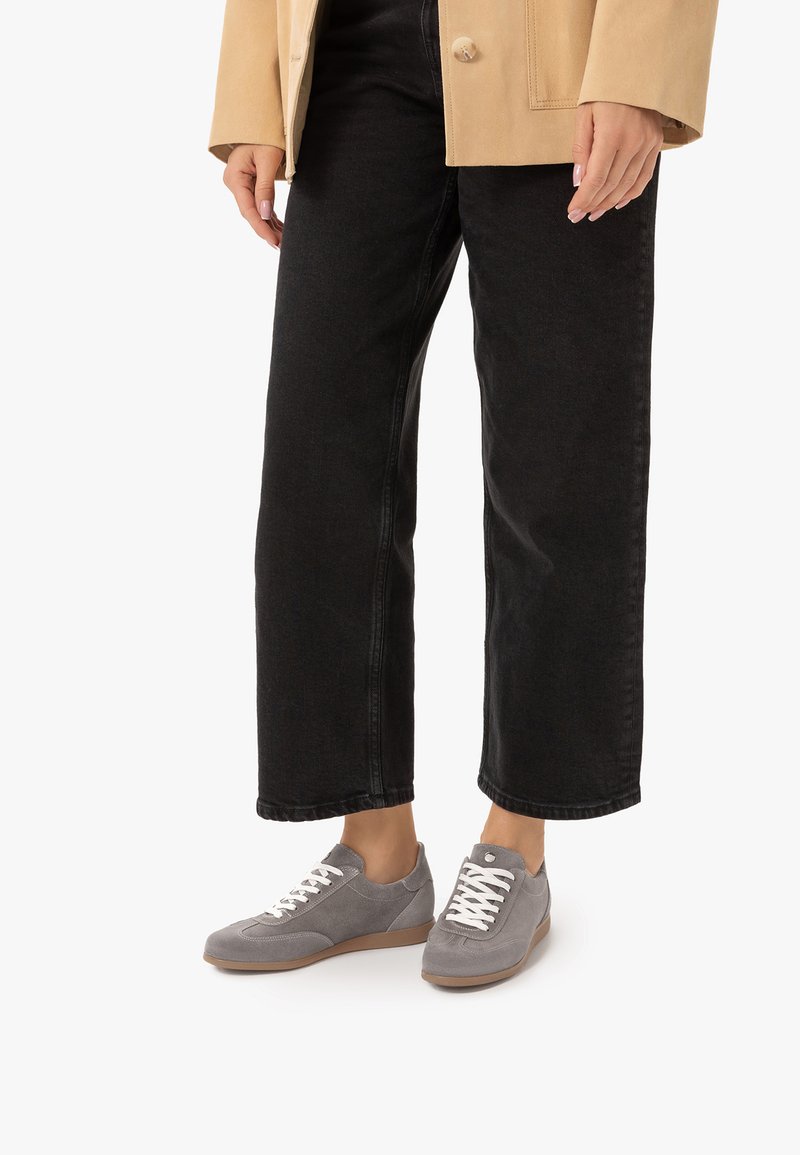 Personne portant un pantalon noir à jambes larges, une veste beige et des baskets grises à lacets avec semelles et lacets blancs sur fond blanc.