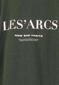 Donkergroen katoenen T-shirt met opvallende witte tekst: "LES'ARCS" en "NEW BAD HABITS" met daaronder een extra kleine tekst. Zachte textuur.